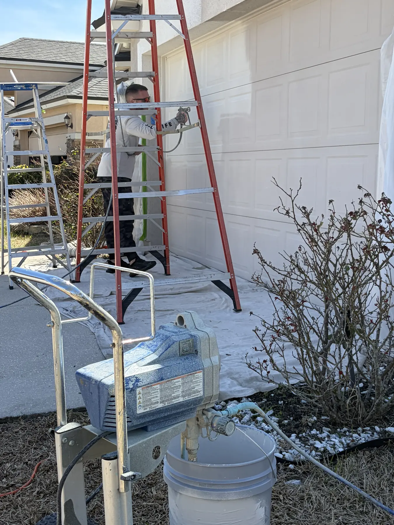 Painter on a ladder spray-painting a home exterior with drop cloths and a paint sprayer set up below